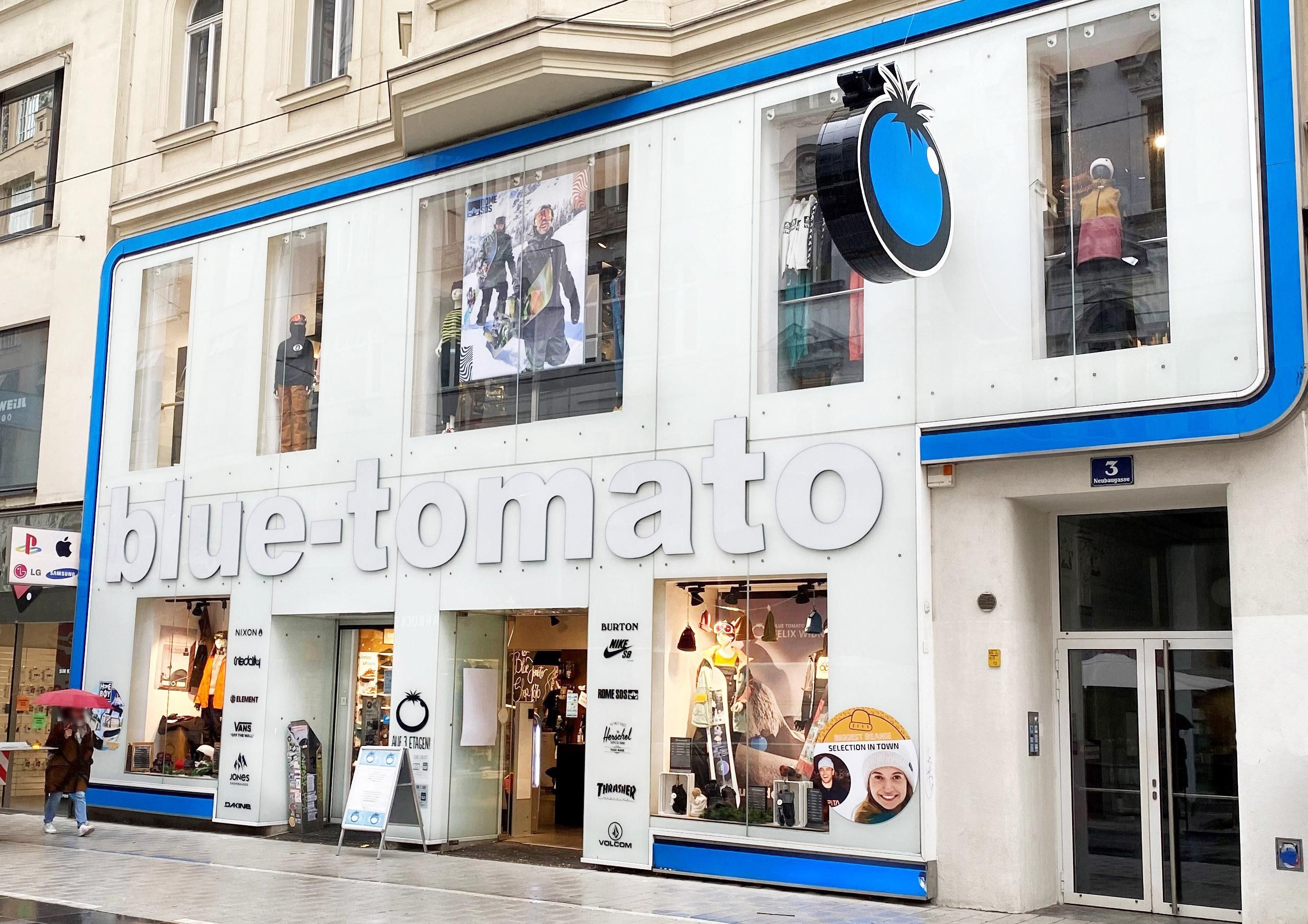 Eine Geschäftsfassade mit ausgestellter Sportkleidung. Die Fassade ist beschriftet mit "blue-tomato" und hat ein Schild von einer blauen Tomate.