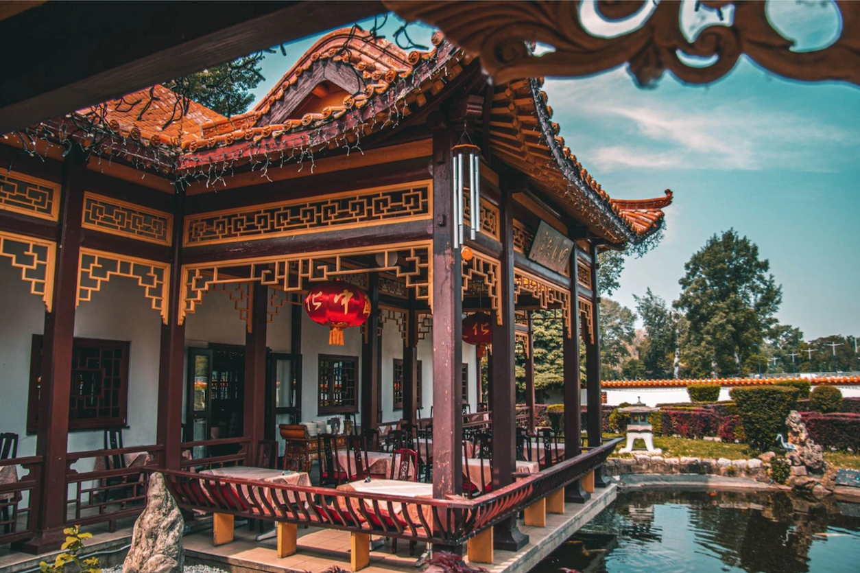 Ein überdachter Außenbereich von einem Restaurant, der einem chinesischen Tempel ähnelt. Unter dem Dach gibt es Tische und Sessel, davor einen Teich und einen Garten im Hintergrund.