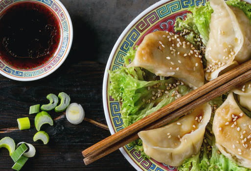 chinesische Teigtaschen und Holzstäbchen in einer Schüssel. Links gibt es eine dunkelrote Soße in einer kleiner Schüssel.