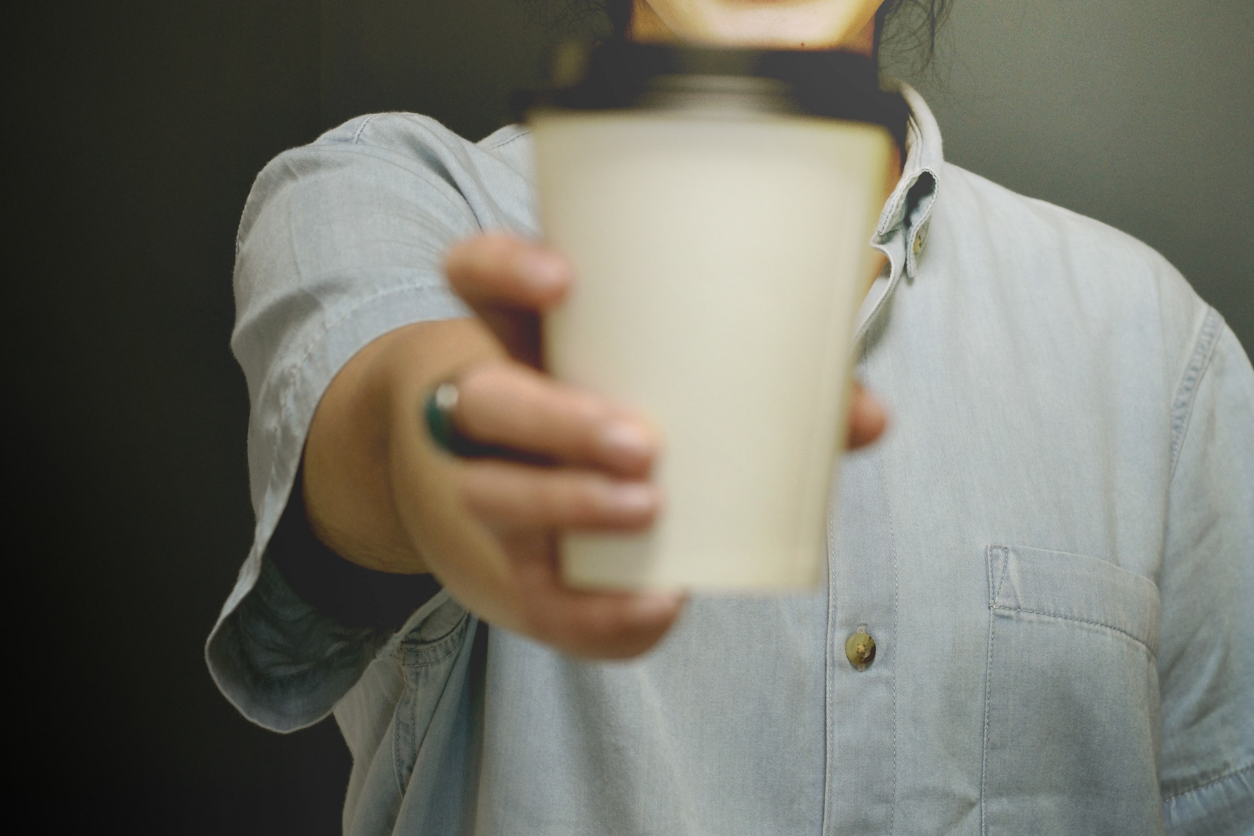 Eine Person hält einen To Go Becher Kaffee in die Kamera