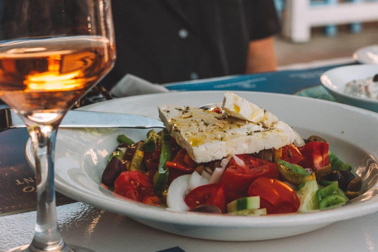 Ein griechischer Salat mit Feta auf einem Teller. Daneben steht ein Glas Wein.