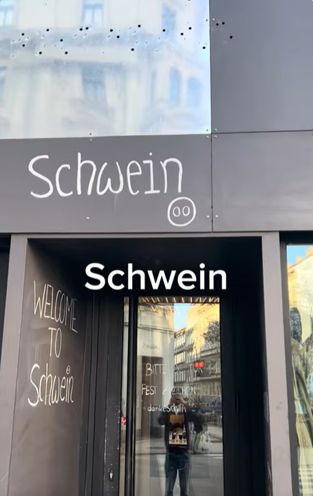 Foto: Außenansicht einer Häuserfronte, auf dessen Fassade Schwein steht. Eine Person spiegelt sich in der Eingangstür und filmt